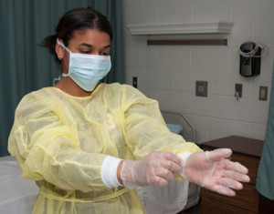 A nursing student demonstrates proper protective gear by wearing a face mask, a gown, and gloves.