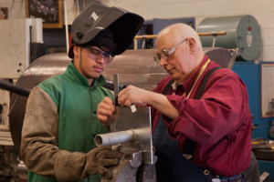 An instructional aide provides assistance to a student working on an assignment in the welding lab.